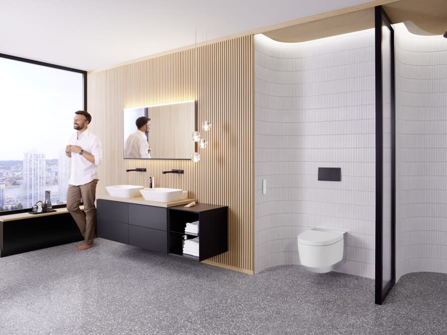 Mężczyzna w przestronnej łazience z ciemnymi meblami i toaletą myjącą Geberit AquaClean Mężczyzna w przestronnej łazience z ciemnymi meblami i toaletą myjącą Geberit AquaClean